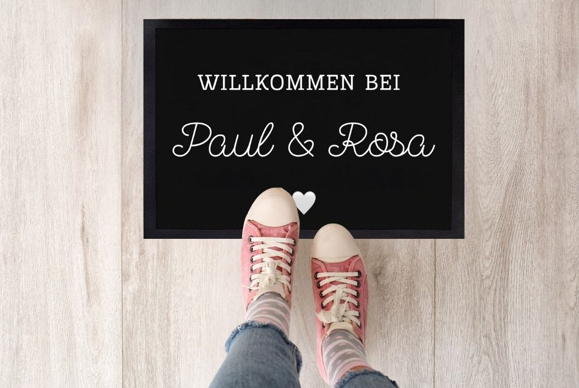 Personalisierte Fußmatte mit der Aufschrift „Willkommen bei Paul & Lisa“ in Weiß auf schwarzem Hintergrund, dekorativ mit Herzsymbol und Holzfußboden